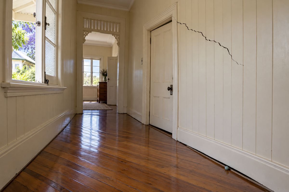 A sloping polished hardwood hallway in an older Toowoomba house with a diagonal crack through the VJ boards over a door, a door planed to fit, a gap opening at the skirting and a marble resting at the low end.